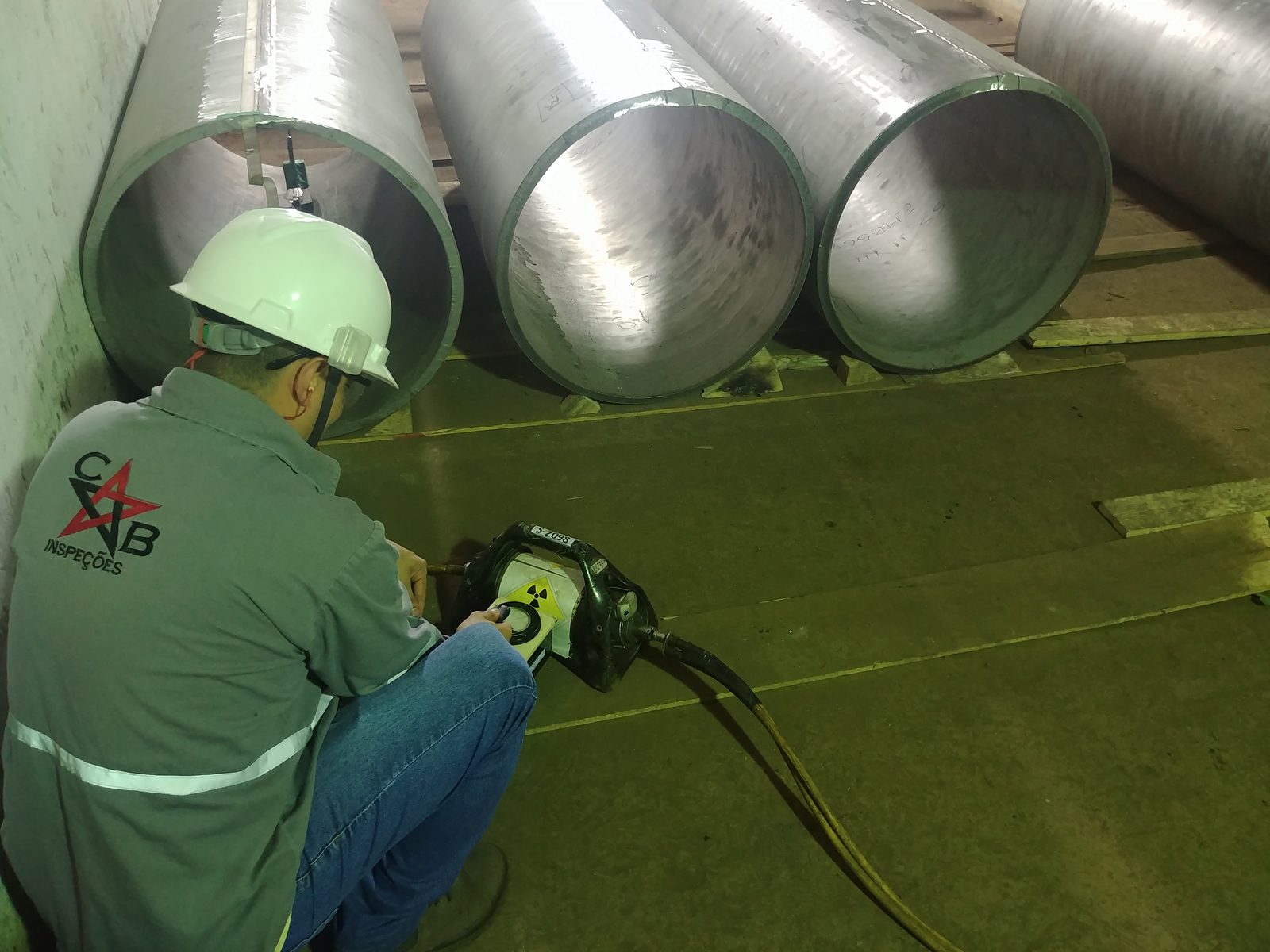 Inspetor de costas, com capacete, operando o equipamento de gamagrafia junto a três tubos de aço inox de grande diâmetro em galpão industrial
