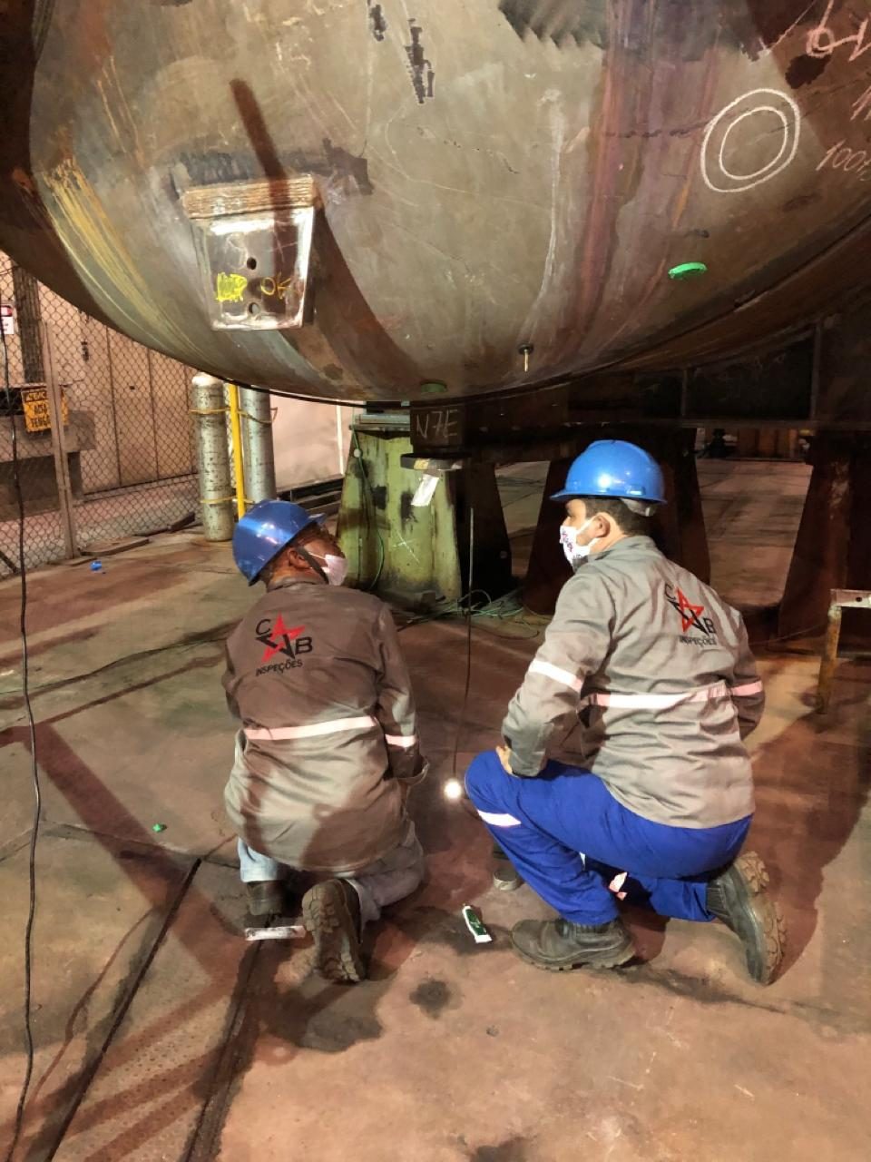 Dois inspetores da CB Inspeções, de capacete e uniforme, preparam o ensaio sob um vaso de pressão na planta do cliente