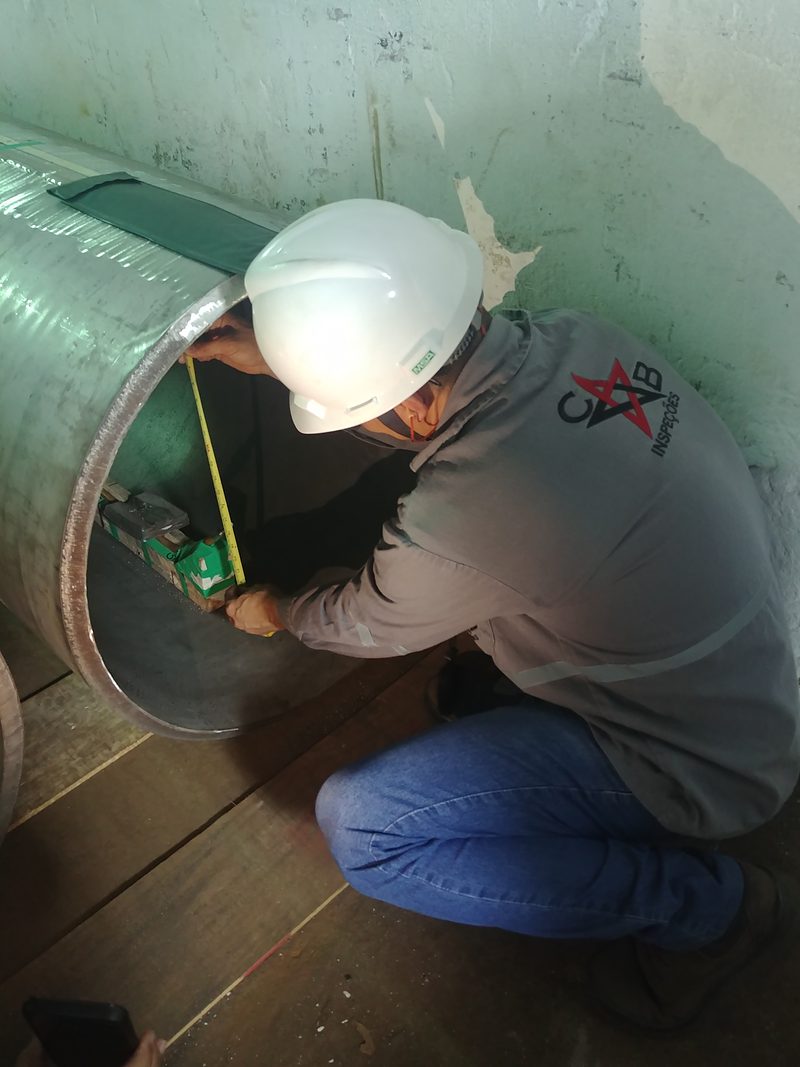 Inspetor da CB Inspeções, de capacete e uniforme, mede com trena a parede de um tubo de grande diâmetro antes do ensaio radiográfico