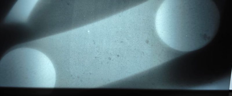 Imagem radiográfica em preto e branco de peça mecânica com dois furos circulares e nervura diagonal